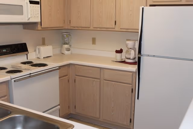 A compact kitchen with light wood cabinets, a white refrigerator, an electric stove with four burners, a microwave above the stove, a toaster, a blender, and a coffee maker on the countertop.