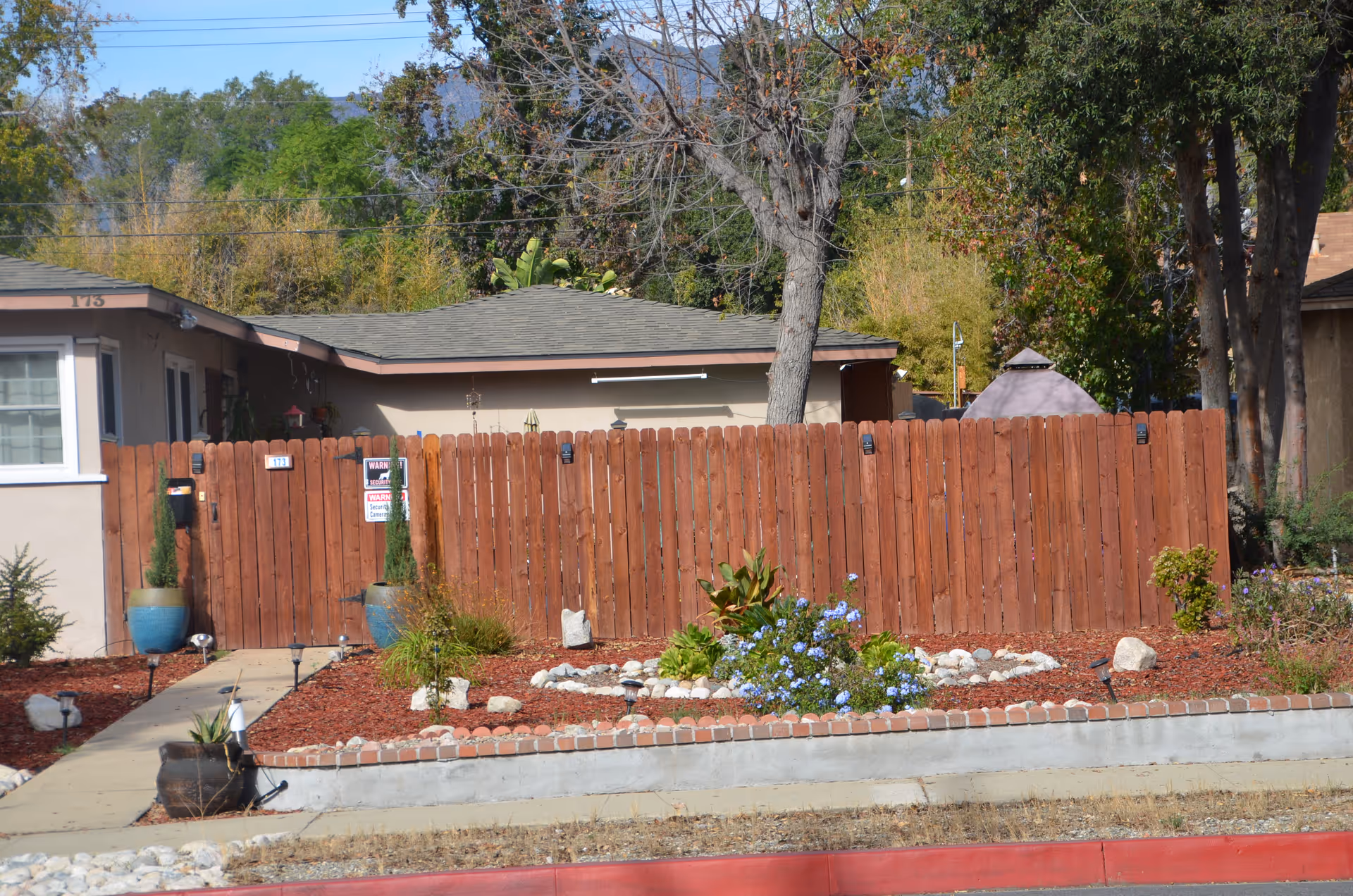 Front yard and wooden fence of a single-story house with potted plants, landscaping, and a tree.