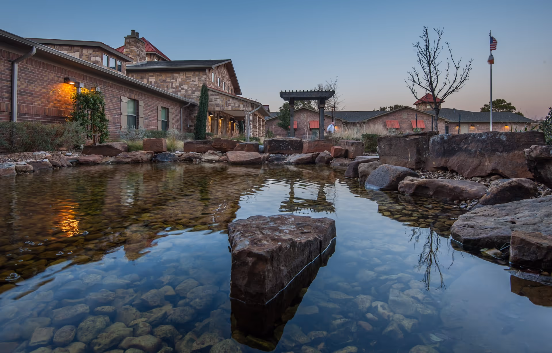 A serene outdoor pond with clear water and large rocks surrounding it, reflecting the nearby buildings and trees. The buildings have a rustic brick and stone exterior with warm lighting, and there is a flagpole with an American flag in the background under a clear sky at dusk.