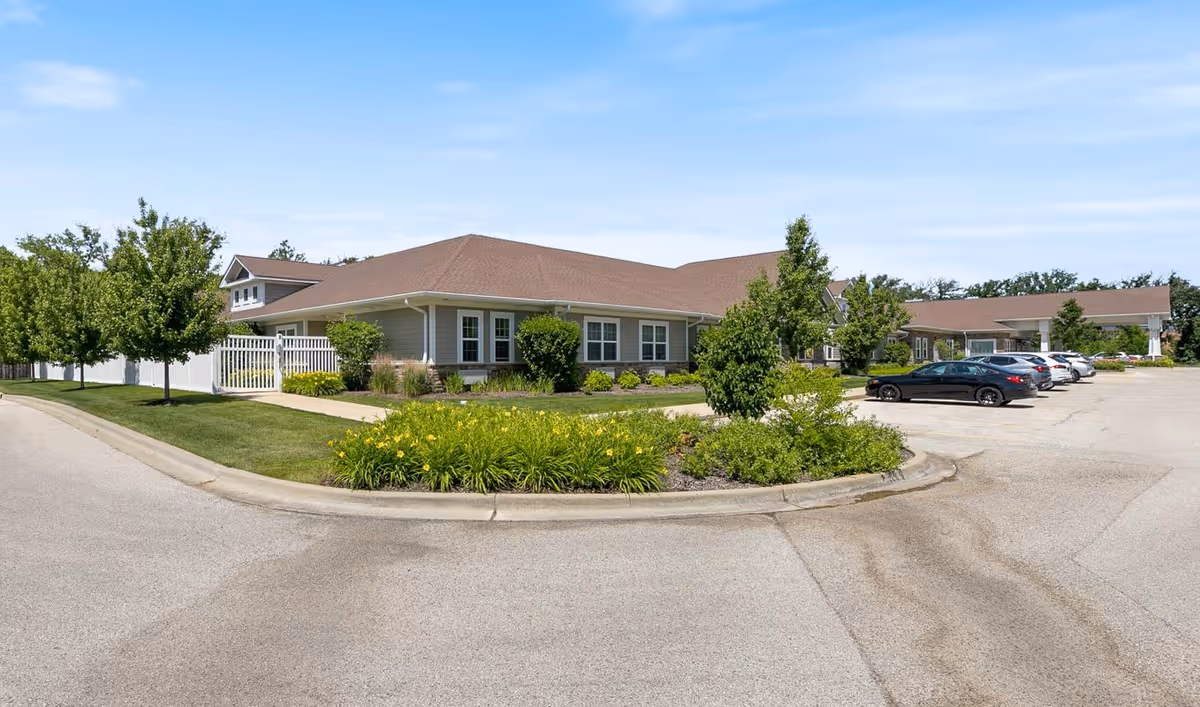 Exterior view of a single-story senior living facility building with a brown roof, beige siding, and multiple windows. The building is surrounded by well-maintained landscaping including green bushes, trees, and flower beds. There is a paved parking lot with several parked cars and a clear blue sky above.