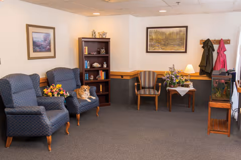 A cozy sitting area in a nursing and rehabilitation center featuring two blue patterned armchairs with a small dog resting on one, a wooden bookshelf with decorative items and books, a wooden chair with striped upholstery, a small table with a lamp and flower arrangement, coat hooks with jackets hanging, framed landscape paintings on the walls, and a fish tank on a wooden stand.