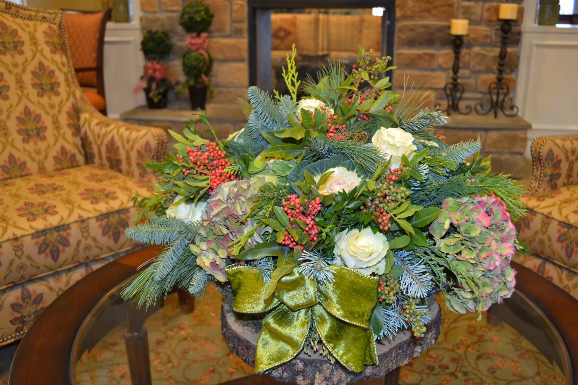 A floral arrangement with white roses, green leaves, red berries, and pine branches tied with a green velvet bow, placed on a glass coffee table in a cozy room with patterned armchairs and a stone fireplace in the background.