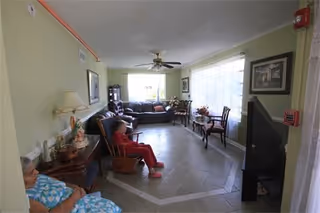 A living room in an adult living facility with two elderly women seated on chairs. The room has light green walls, a ceiling fan, a large window with sheer curtains, a dark brown sofa, and a television on a stand. There are framed pictures on the walls and a small table with flowers near the window.