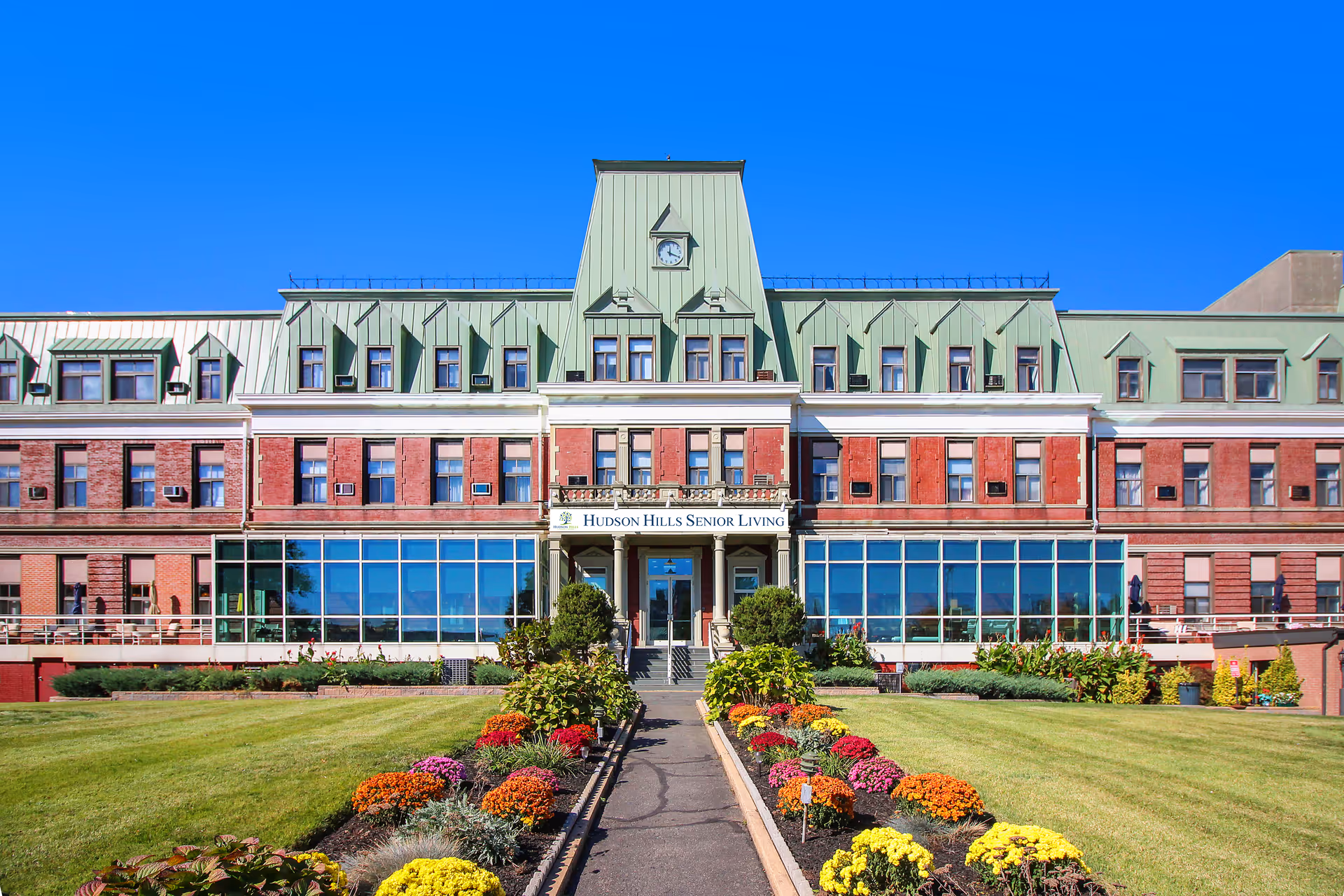 Front exterior view of a large senior living facility building with a green roof and red brick walls. The building has multiple windows and a clock tower in the center. A well-maintained garden with colorful flowers and a paved walkway leads up to the entrance. The sign above the entrance reads 'Hudson Hills Senior Living'.