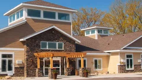 Exterior view of Woodstone Senior Living facility showing a modern building with beige siding, stone accents, large windows, and a wooden pergola over the entrance. Trees with autumn foliage are visible in the background under a clear blue sky.