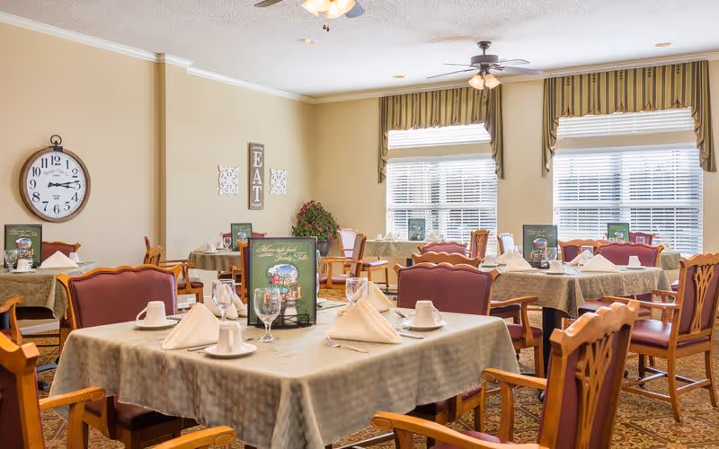 A dining room with several tables covered in beige tablecloths, each set with white napkins, cups, glasses, and menus. The room has large windows with striped valances, a wall clock, and decorative wall art including the word 'EAT'. The chairs have wooden frames with red cushioned seats and backs.