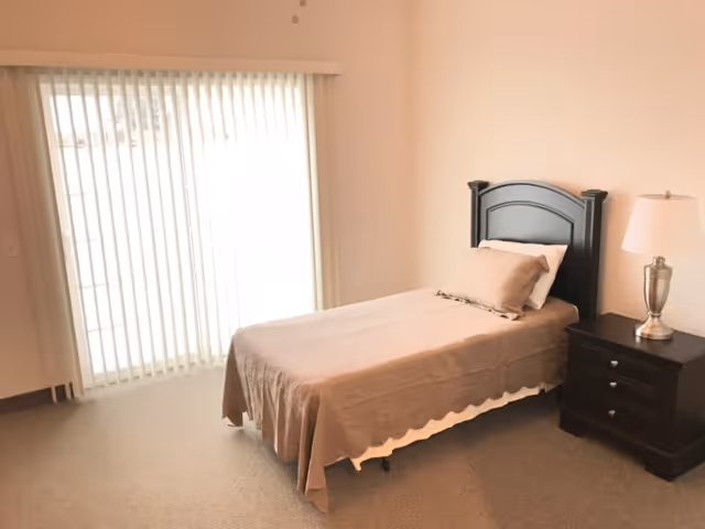 A simple bedroom with a single bed covered in a beige bedspread and a matching pillow. Next to the bed is a dark wooden nightstand with three drawers and a silver lamp with a white lampshade. The room has beige walls and carpeted floor, and a large window with vertical blinds letting in natural light.