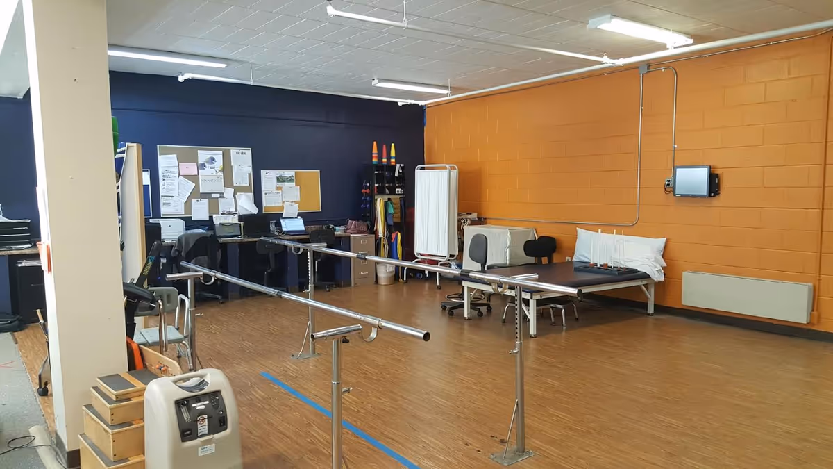 A rehabilitation room with parallel bars for walking exercises, a treatment table with pillows, office desks with computers, chairs, and a bulletin board on the wall. The room has an orange and dark blue painted brick wall and wood-patterned flooring.