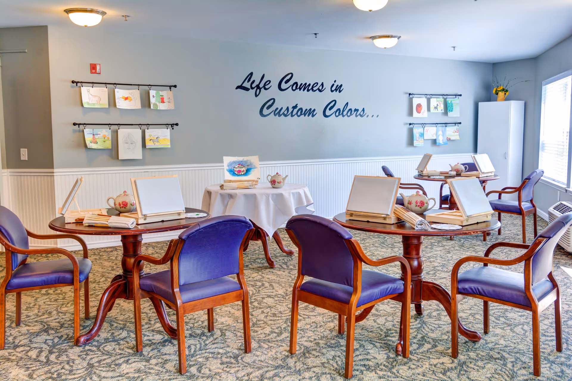 A bright and inviting activity room with round wooden tables and purple cushioned chairs arranged around them. Each table has blank canvases on easels, paintbrushes, and teapots, suggesting a painting or art activity setup. The walls are light blue with white wainscoting, and children's artwork is displayed on hanging rods. On the wall, there is a decorative phrase that reads 'Life Comes in Custom Colors...'. Large windows allow natural light to fill the room.