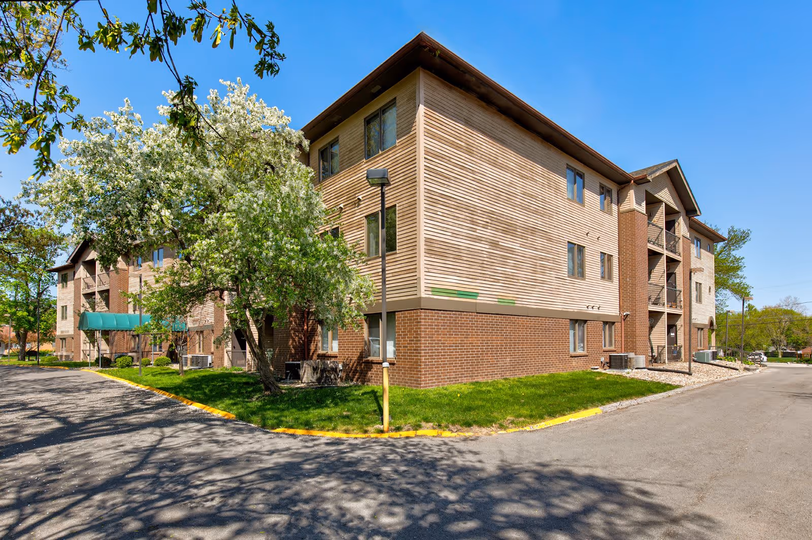 Three-story brick-and-siding senior apartment building with trees, lawn, and a driveway under a clear blue sky.