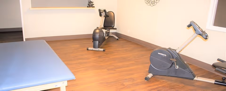 A room with wooden flooring containing two stationary exercise bikes and a padded treatment or massage table against a white wall with a small window and decorative wall art.