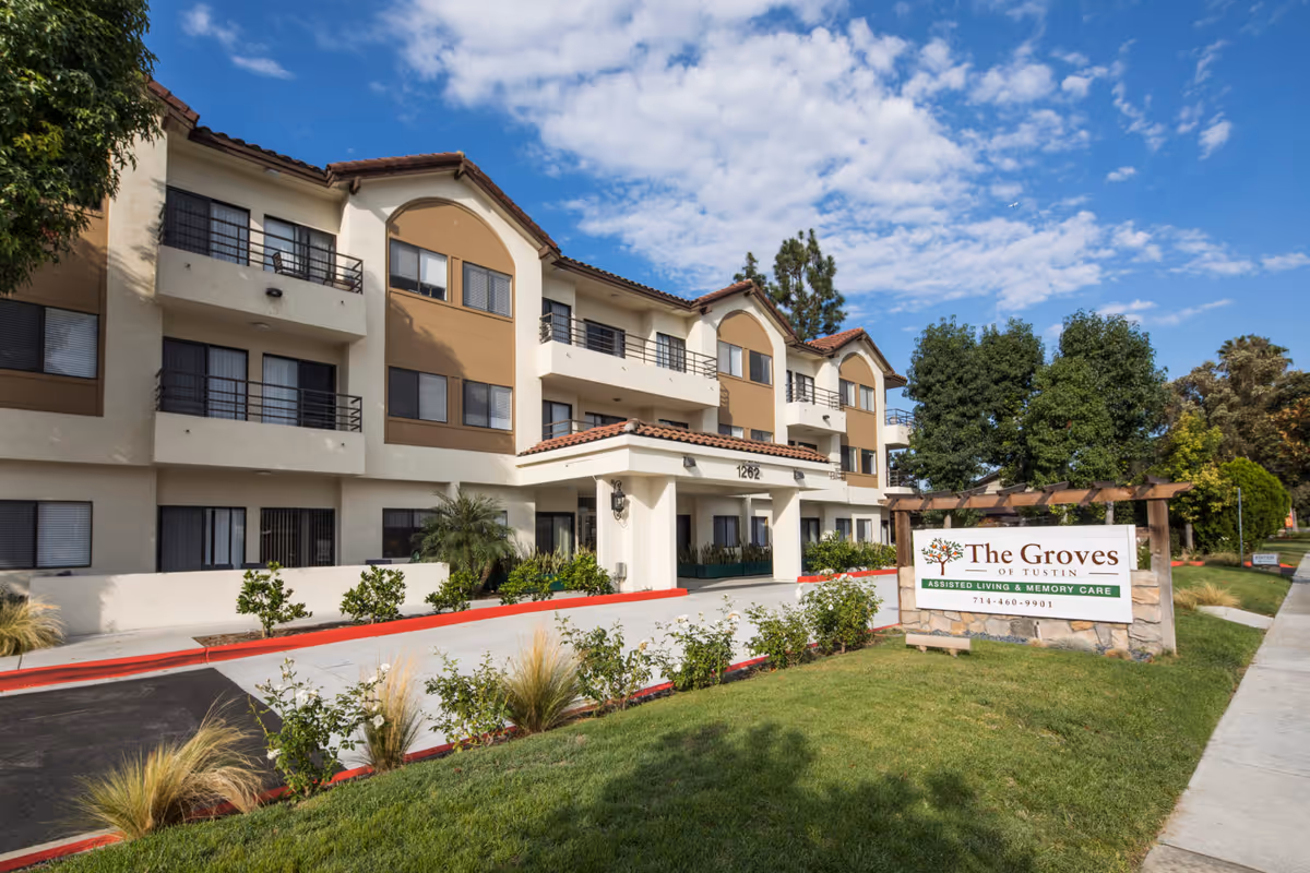 Three-story beige assisted living building with balconies, landscaped lawn, and an entrance sign reading 'The Groves' under a blue sky.