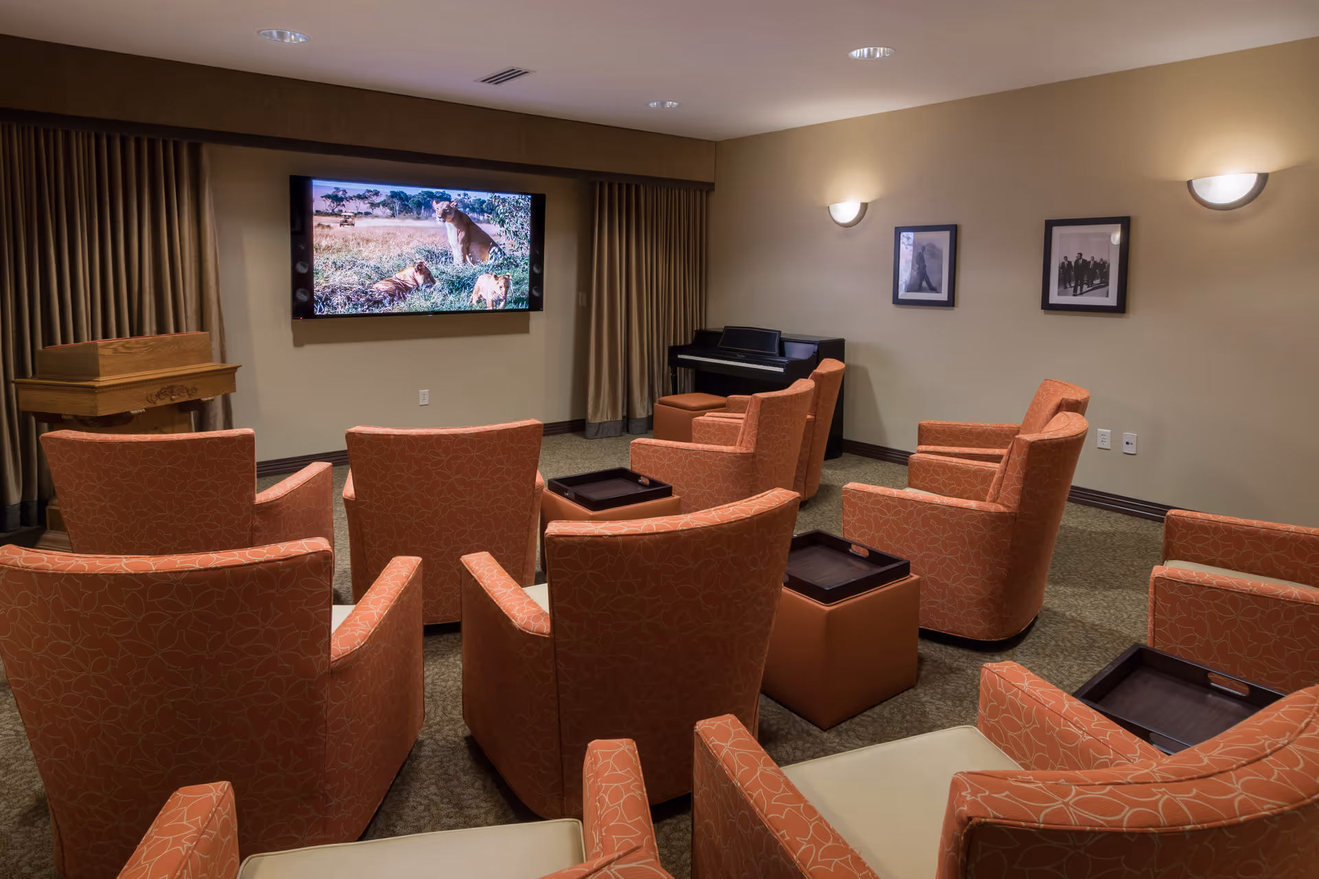A cozy media room with several orange patterned armchairs arranged facing a wall-mounted flat screen TV displaying a nature scene with lions. The room has beige walls, two framed black and white photographs, a piano in the corner, and soft lighting from wall sconces.