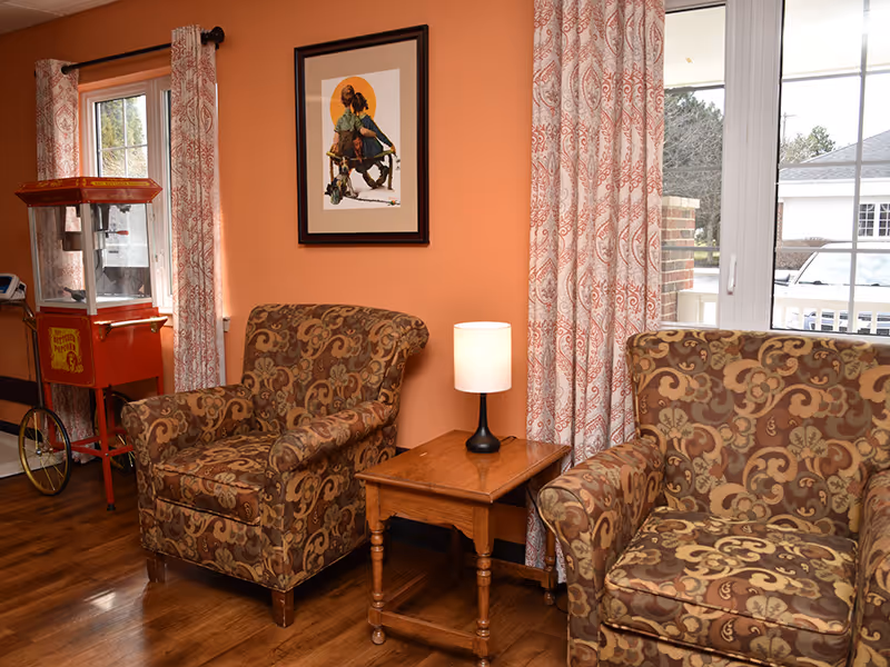 A cozy sitting area with two patterned armchairs separated by a wooden side table with a small lamp. The walls are painted orange and decorated with a framed artwork. There are patterned curtains on the windows, and a vintage-style popcorn machine is visible in the corner.