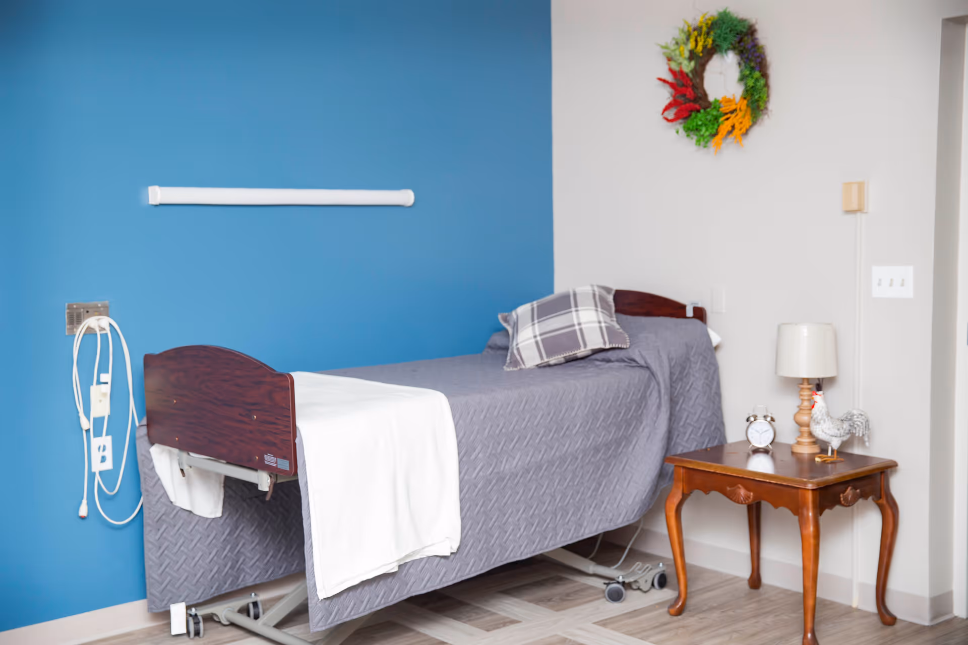 A single hospital-style bed with a gray quilted blanket and a plaid pillow is positioned against a blue and white wall. A white towel is draped over the foot of the bed. Next to the bed is a wooden side table with a small lamp, an alarm clock, and a decorative rooster figurine. A colorful wreath hangs on the white wall above the table.