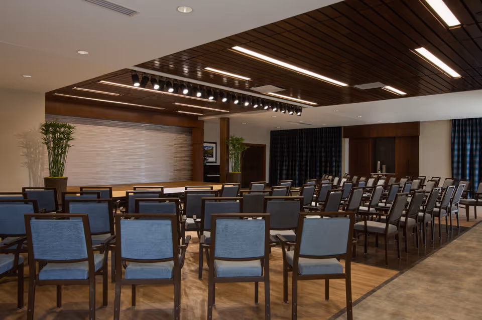 A large multipurpose meeting room with rows of blue-upholstered chairs facing a stage under recessed and track lighting.