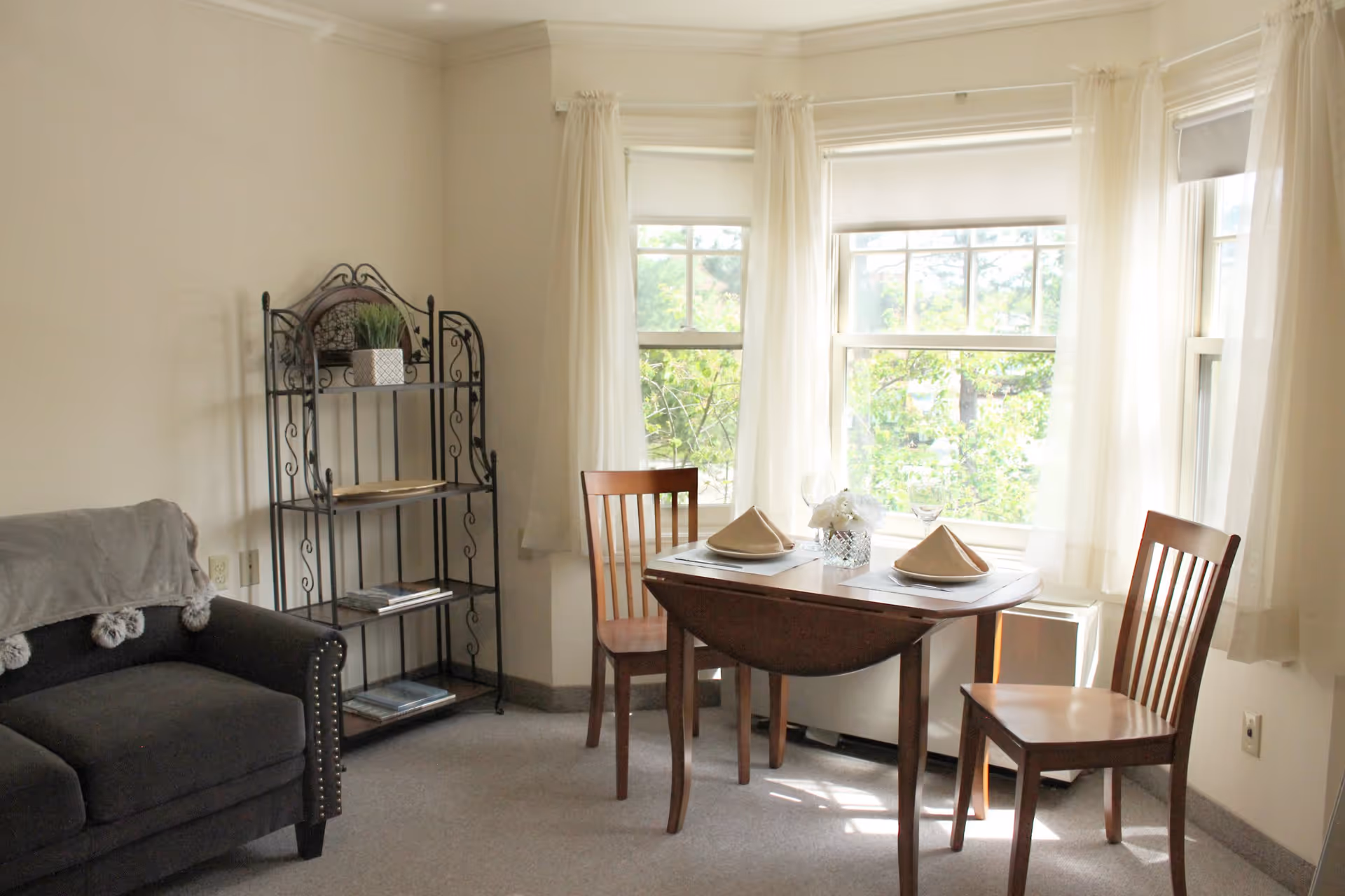 A cozy living area with a dark gray armchair draped with a light gray blanket, a black metal shelving unit with decorative items, and a small wooden dining table set for two with folded napkins and a small flower arrangement. The room has large windows with sheer white curtains allowing natural light to brighten the space.
