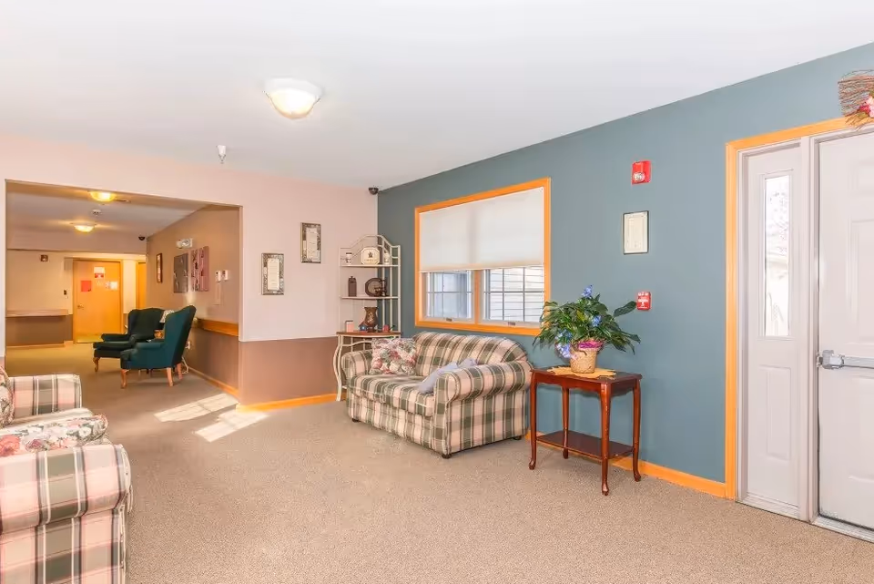 A cozy seating area in a senior living facility with plaid upholstered sofas, a small wooden table with a potted plant, and a shelving unit with decorative items. The walls are painted in muted tones with a window letting in natural light. In the background, there are two green armchairs and a hallway leading to other rooms.