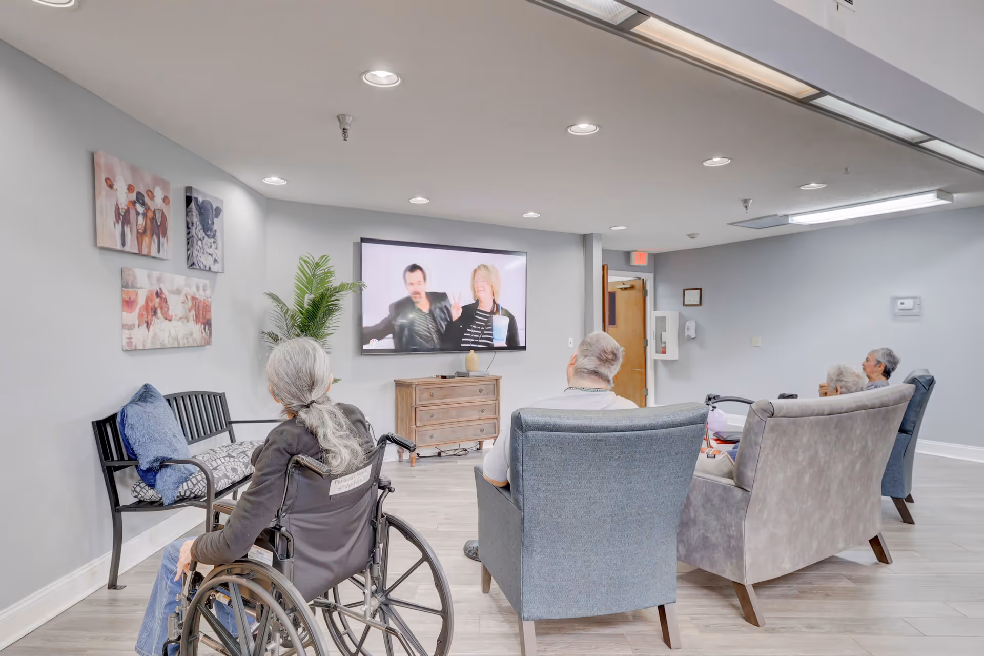 A group of elderly people sitting in a common room watching a television mounted on the wall. One person is in a wheelchair, and others are seated on armchairs and a loveseat. The room has light gray walls, wood flooring, and animal-themed artwork on the wall. A wooden cabinet is placed under the TV, and a green plant is in the corner.