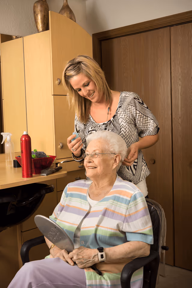 An elderly woman with white hair and glasses sits in a chair holding a hand mirror, smiling as a younger woman styles her hair in a room with wooden cabinets and a counter with hair care products.