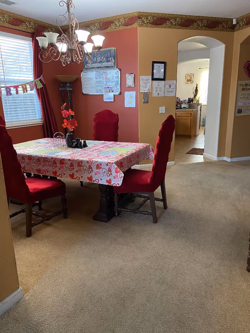 A dining room with a rectangular table covered with a heart-patterned tablecloth. Four red cushioned chairs surround the table. A chandelier with multiple lights hangs above the table. There is a window with blinds and red curtains on the left side, and a birthday banner hanging across the window. The walls are painted in warm tones of red and beige with a decorative border near the ceiling. Various papers and a whiteboard are pinned to the wall. An open doorway leads to a kitchen area in the background.