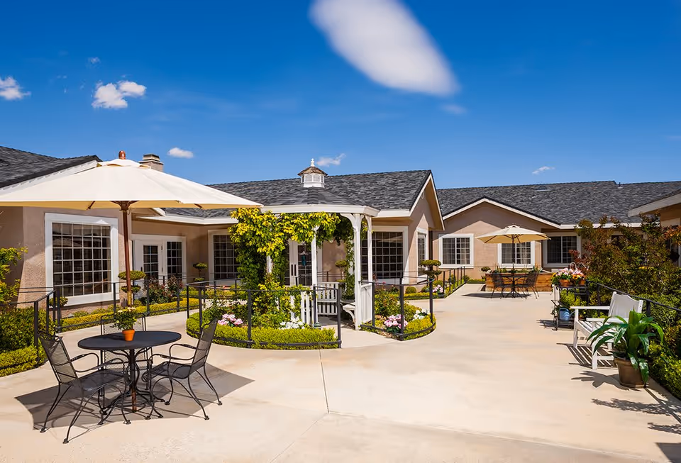 Outdoor courtyard area of Sterling Commons Memory Care Community featuring a paved walkway, a white gazebo with greenery, patio tables with umbrellas, chairs, and potted plants under a clear blue sky.
