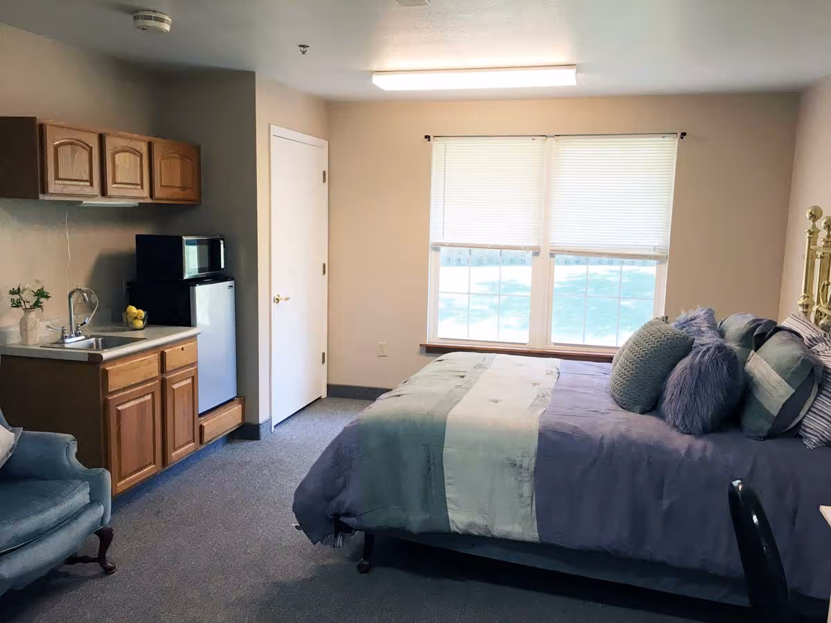 A cozy bedroom with a bed covered in blue and gray bedding and multiple pillows. There is a window with blinds letting in natural light. To the left, there is a small kitchenette area with a sink, wooden cabinets, a mini fridge, and a microwave. A blue upholstered chair is partially visible in the corner.