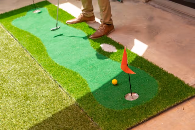 A person standing on an indoor putting green mat holding a golf putter, aiming at a hole with a red flag and a yellow golf ball nearby.