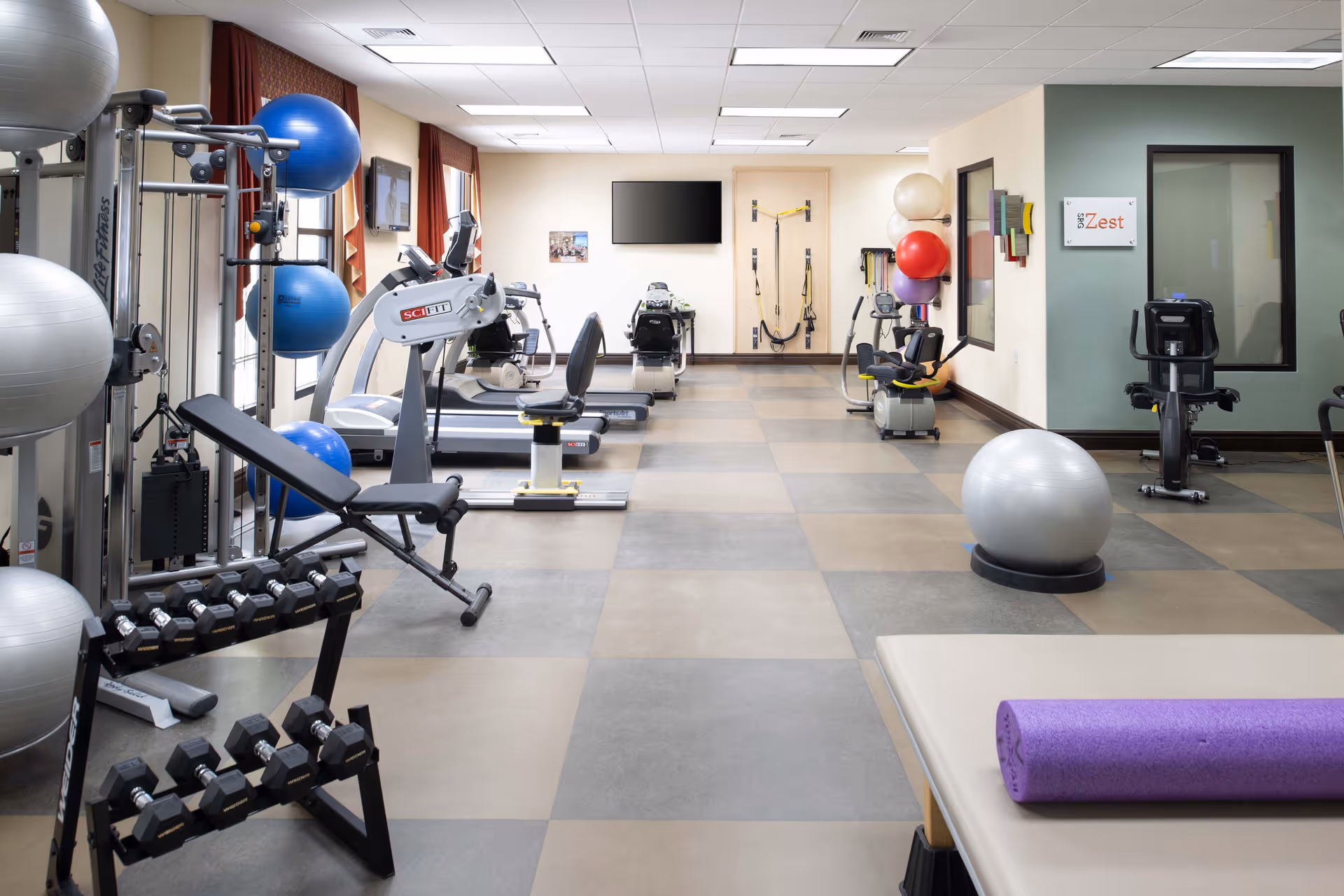 A well-lit fitness room with various exercise equipment including dumbbells, exercise balls, stationary bikes, treadmills, and resistance training machines. The room has large windows with red curtains, a wall-mounted TV, and a sign that reads 'Zest'.