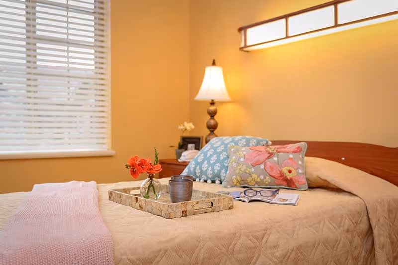 A cozy bedroom with a neatly made bed featuring a beige quilt and decorative pillows with floral patterns. A wooden tray on the bed holds a small vase with red flowers and a cup. A table lamp is lit on a nightstand beside the bed, and a window with white blinds lets in natural light.