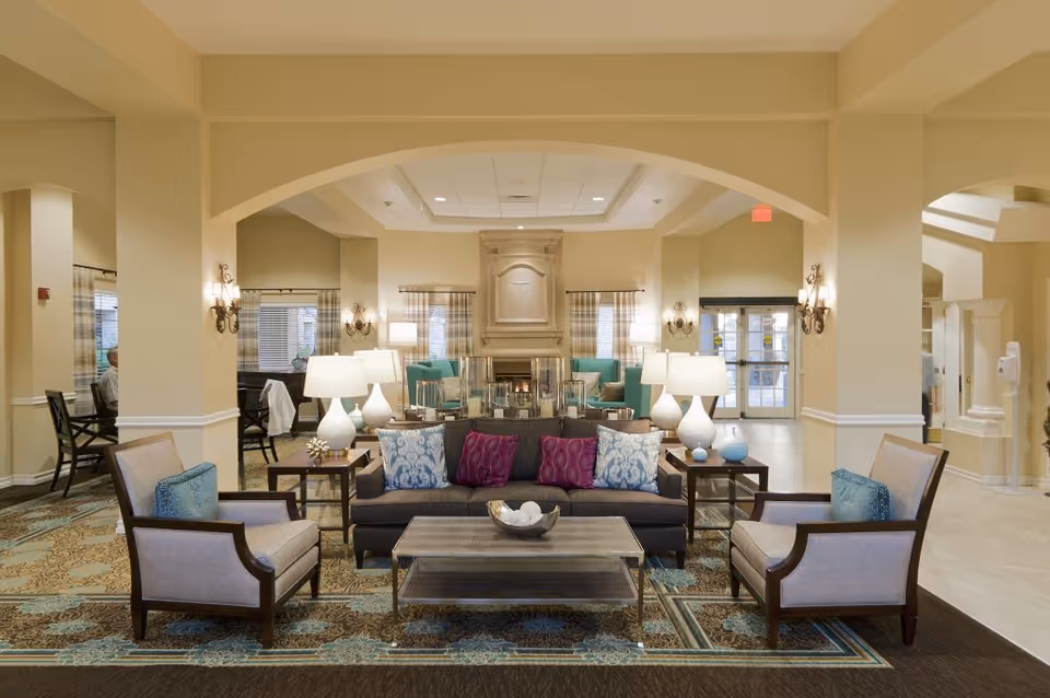 A spacious and well-lit living room area in a senior living facility with a central dark gray sofa adorned with blue and purple patterned pillows, flanked by two beige armchairs with blue cushions. The room features a large decorative fireplace in the background, multiple table lamps on side tables, wall sconces, and large windows with plaid curtains. The floor has a patterned carpet, and there are archways leading to other parts of the facility.