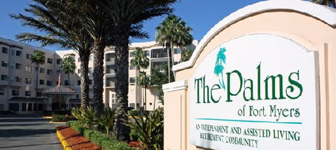 Exterior view of The Palms of Fort Myers senior living facility with a large sign in the foreground and a multi-story building with palm trees and landscaping in the background.