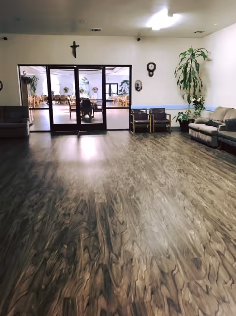 Spacious senior living common room with wood-patterned flooring, sofas and chairs, potted plants, and glass doors opening to a dining/dining area.