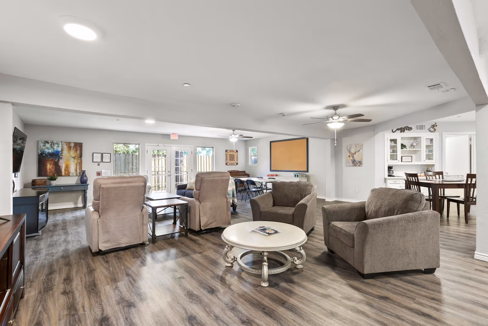 A spacious and well-lit common living area in an assisted living facility featuring comfortable armchairs and recliners arranged around a round coffee table. The room has wood-style flooring, ceiling fans, and large windows with glass doors leading outside. In the background, there is a dining area with a table and chairs, a bulletin board on the wall, and decorative artwork.