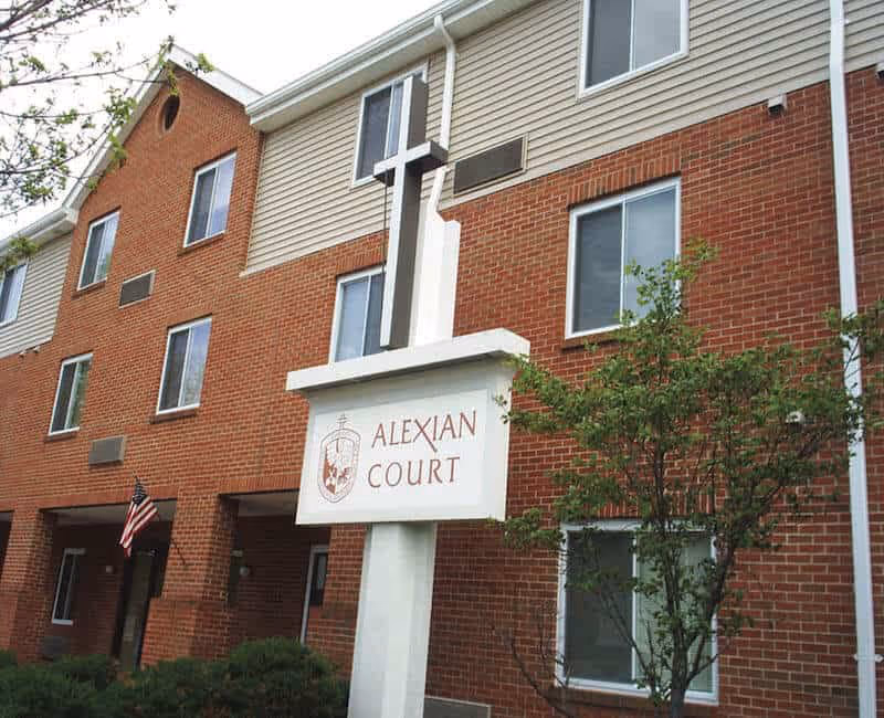 Exterior view of a brick and siding building with multiple windows and an American flag near the entrance. In front of the building is a white signpost with a cross on top displaying the name 'Alexian Court' along with a crest.