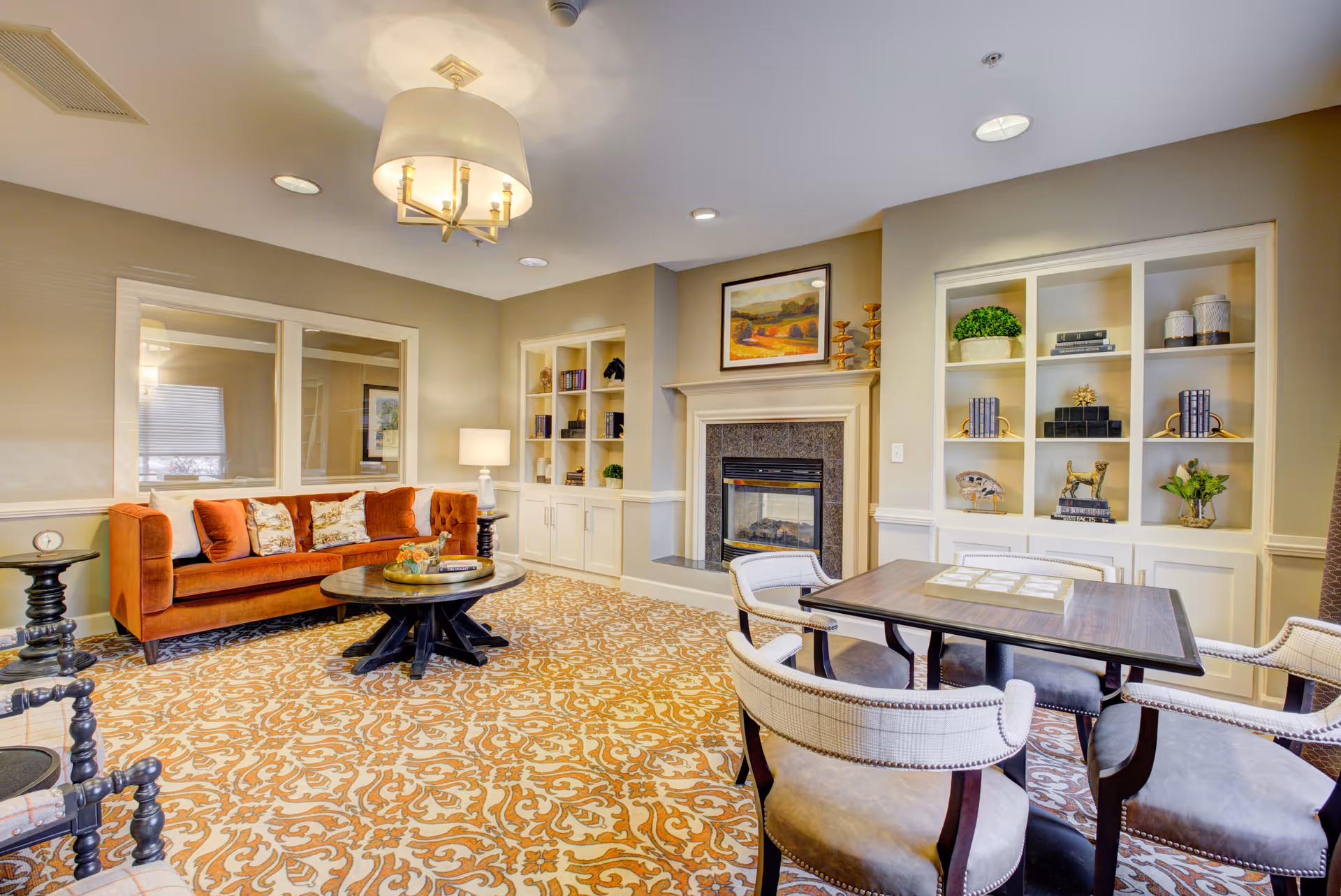 A cozy living room area with a burnt orange sofa adorned with patterned pillows, a round wooden coffee table, and a patterned carpet. There is a fireplace with a framed landscape painting above it, built-in shelves with decorative items, and a square table surrounded by four upholstered chairs. The room is softly lit by a ceiling light fixture and a table lamp.
