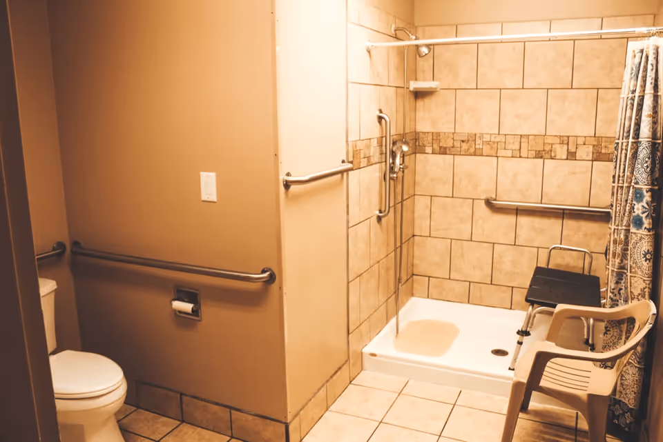 A bathroom with beige tiled walls and floor, featuring a toilet with grab bars on the walls, a walk-in shower with a shower chair, grab bars, and a patterned shower curtain. There is also a plastic chair placed near the shower.