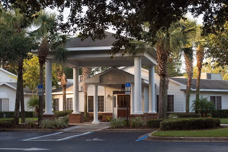 Entrance of a senior living facility with a covered drop-off area supported by white columns, surrounded by palm trees and landscaping. The building is single-story with white siding and multiple windows. Handicap parking spaces are visible in front.