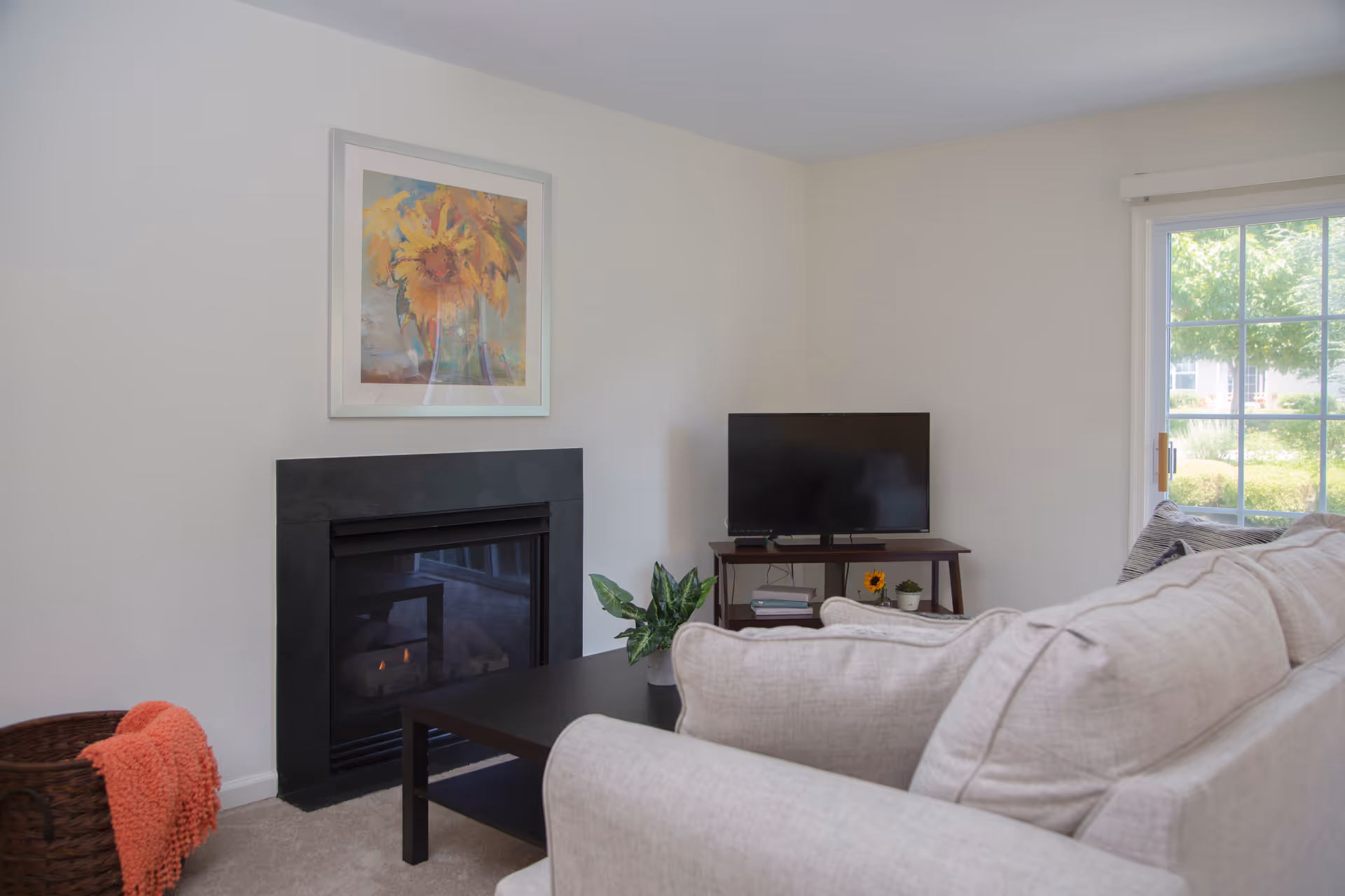A cozy living room with a beige sectional sofa, a black coffee table with a small green plant, a flat-screen TV on a wooden stand, a fireplace with a black surround, and a framed painting of sunflowers above the fireplace. A large window lets in natural light and shows greenery outside.