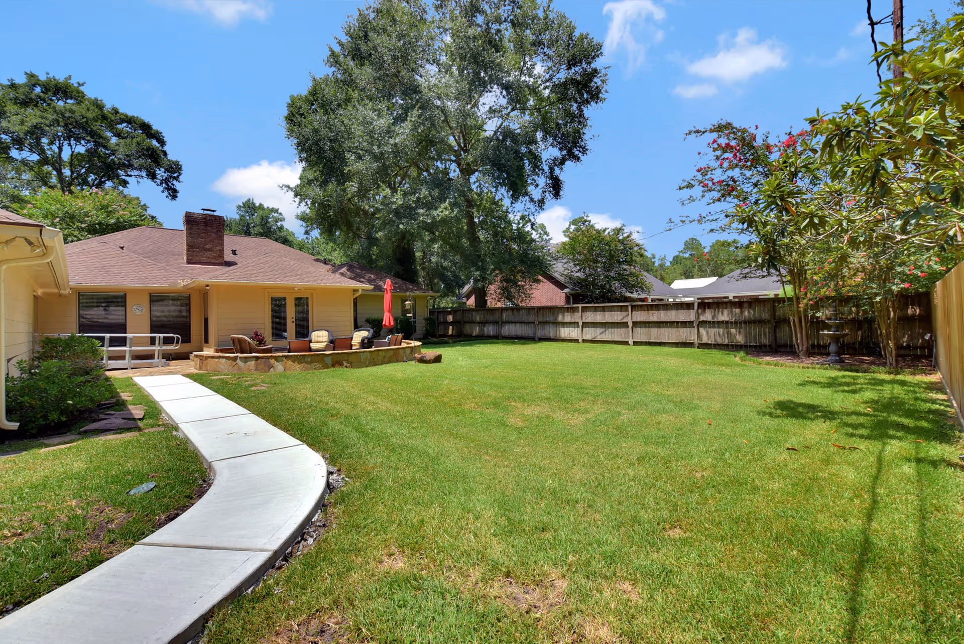 Spacious backyard with a curved concrete walkway leading to a patio and single-story house under a blue sky.