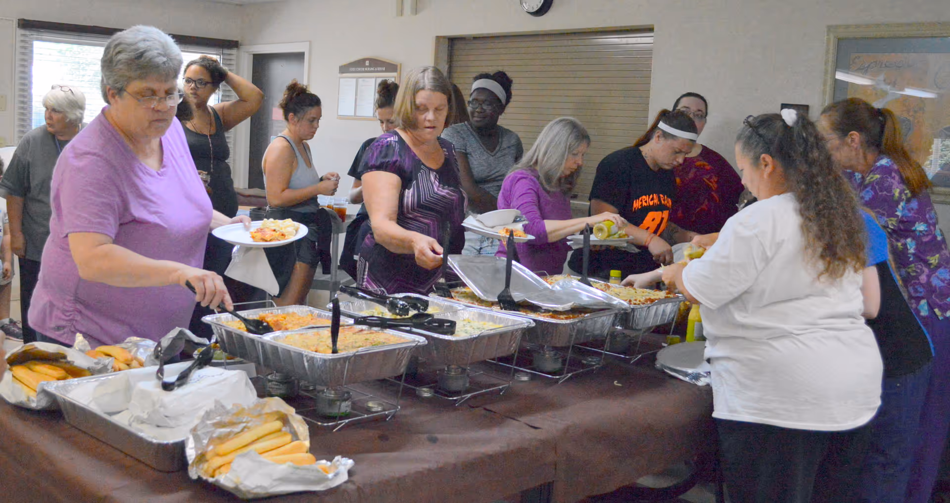 A group of people serving themselves food from a buffet table with trays of hot dishes and bread rolls in a communal dining area.