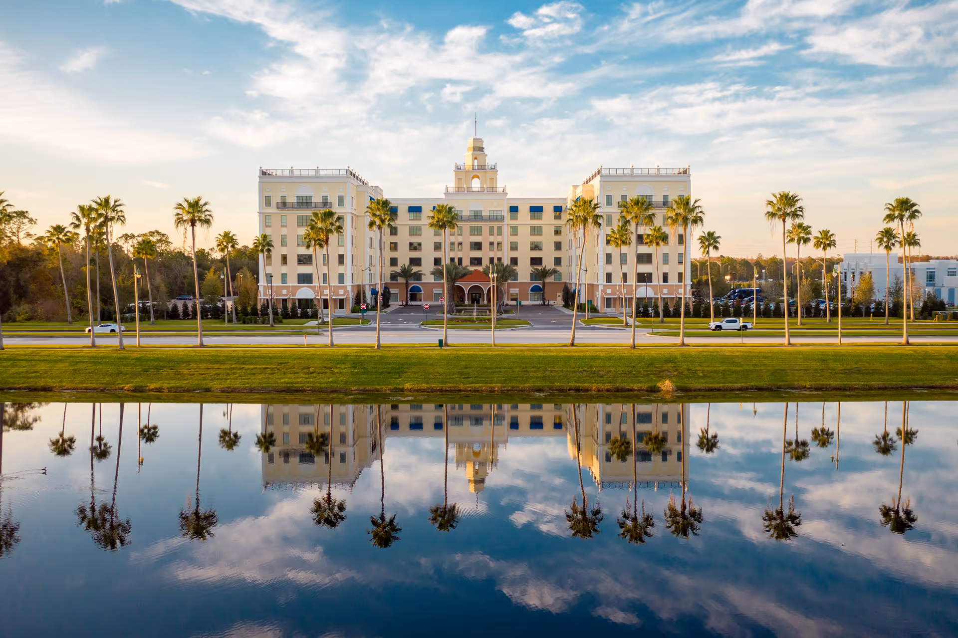 A large multi-story building with a central tower, surrounded by palm trees and reflected in a calm body of water in the foreground under a partly cloudy sky.