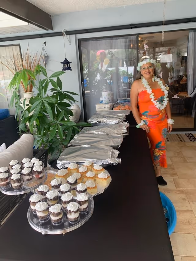A woman dressed in a bright orange floral dress and white flower lei stands next to a table covered with a black tablecloth. The table holds several trays covered with foil, plates of desserts, and small dessert cups topped with whipped cream. The setting appears to be an indoor-outdoor area with plants and sliding glass doors in the background.