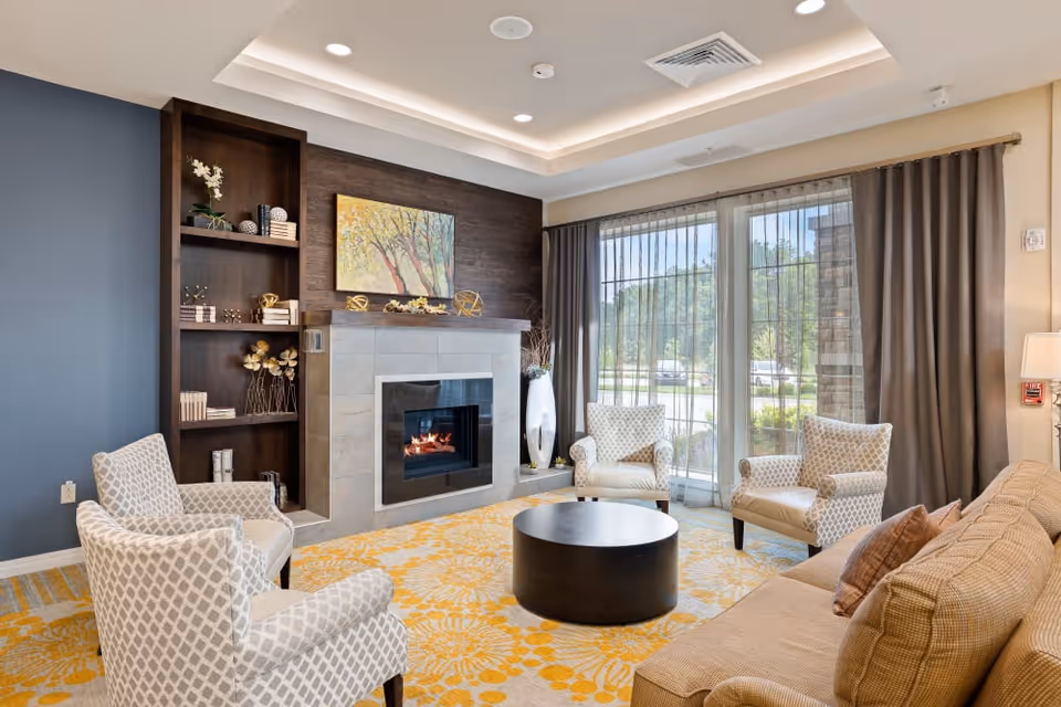A cozy living room with a modern fireplace set in a tiled wall with wooden shelves on the left. The room features a beige sofa with brown pillows, three patterned armchairs, a round black coffee table, and a yellow floral carpet. Large windows with sheer and dark curtains allow natural light to fill the space, showing a view of greenery outside.