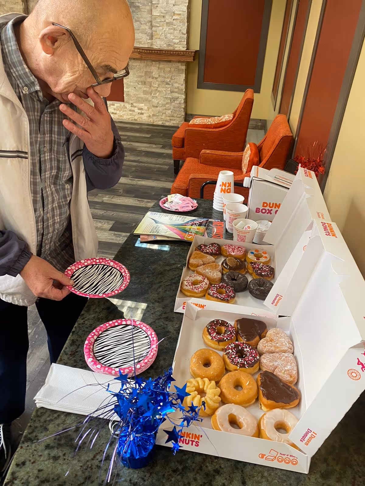 An elderly man wearing glasses and a jacket is standing at a counter with two open boxes of assorted Dunkin' Donuts donuts. There are two pink and black zebra-striped paper plates on the counter, along with napkins, cups, and a small blue star-shaped decoration. In the background, there are orange armchairs and a stone fireplace.