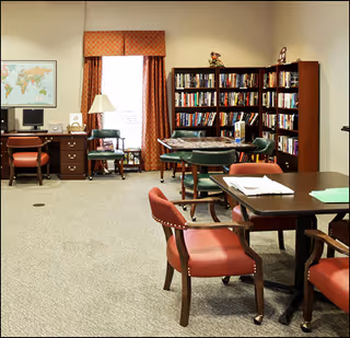 A spacious interior common room with tables and red-cushioned chairs, bookshelves, a computer desk, and a curtained window.