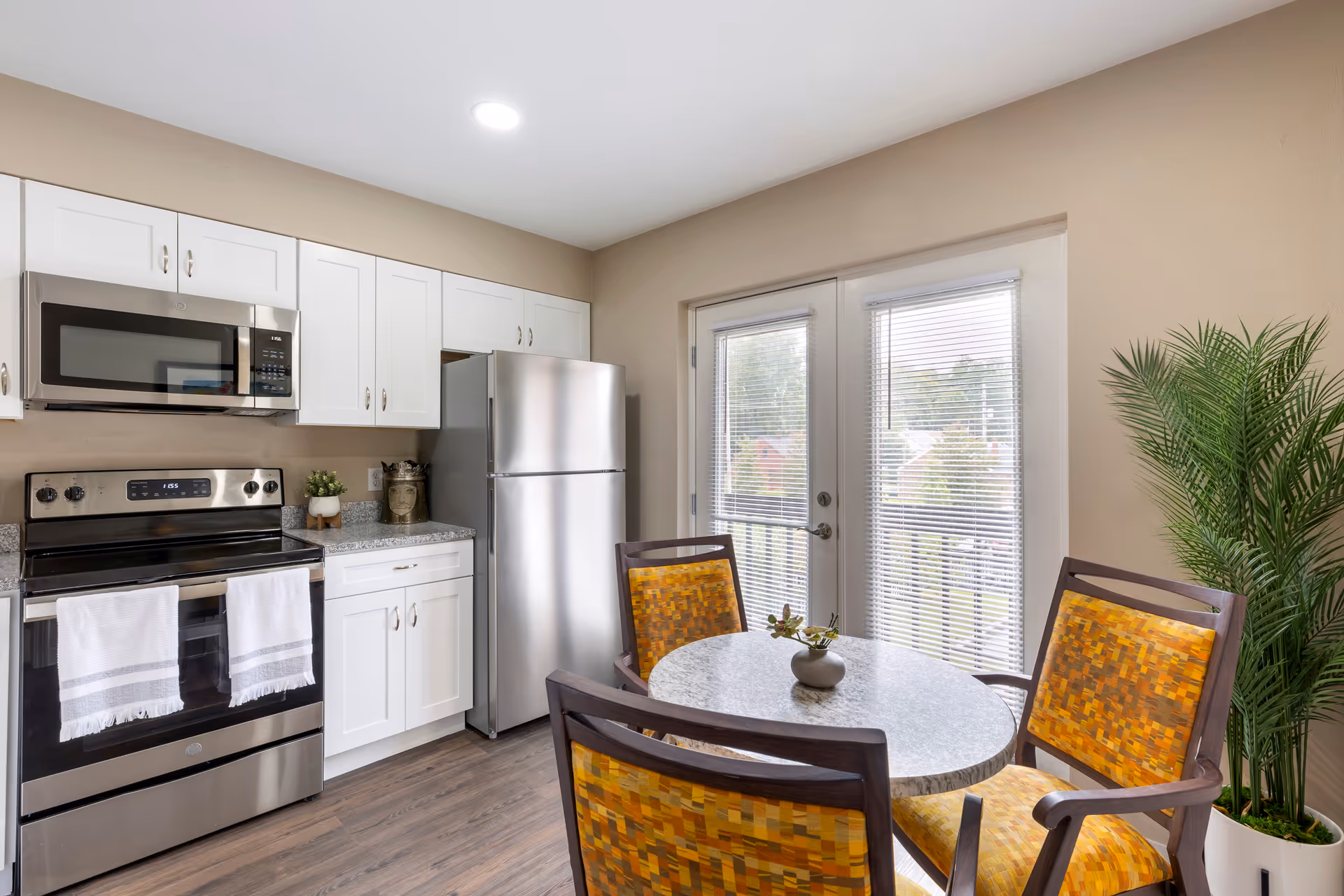 A bright kitchen area with white cabinets, stainless steel appliances including a microwave, stove, and refrigerator. There is a round dining table with four wooden chairs that have yellow patterned cushions. A large potted plant is placed near the glass doors with blinds, leading to an outdoor view.