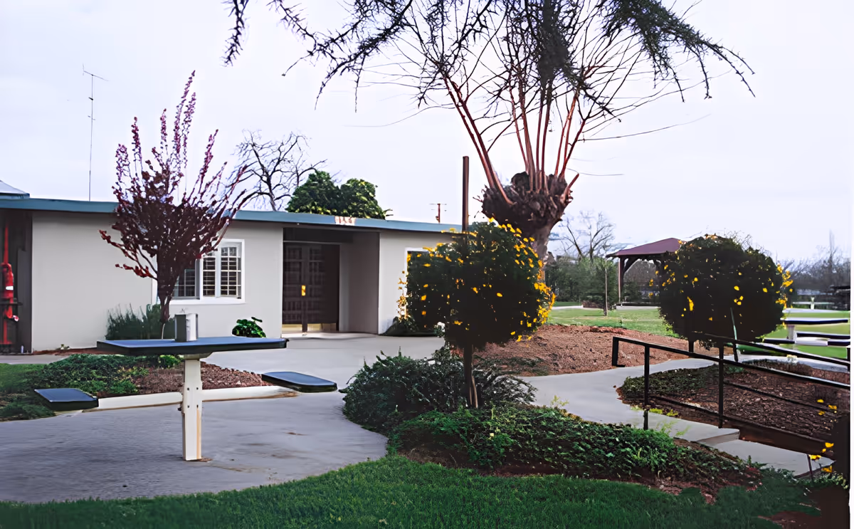 Outdoor area of Davis Guest Home 8 featuring a concrete pathway, green grass, trimmed bushes with yellow flowers, a small tree with bare branches, a picnic table with attached benches, and a single-story building with a window and entrance door in the background.