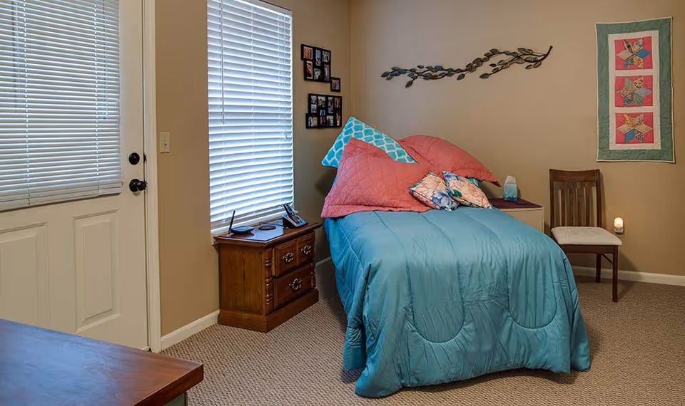 A cozy bedroom with a single bed covered in a teal comforter and multiple pillows in pink and floral patterns. There is a wooden nightstand with a phone and other small items next to the bed. The room has beige walls with a decorative metal wall art and a quilt hanging. A wooden chair with a cushion is placed against the wall, and there is a door and window with closed blinds.