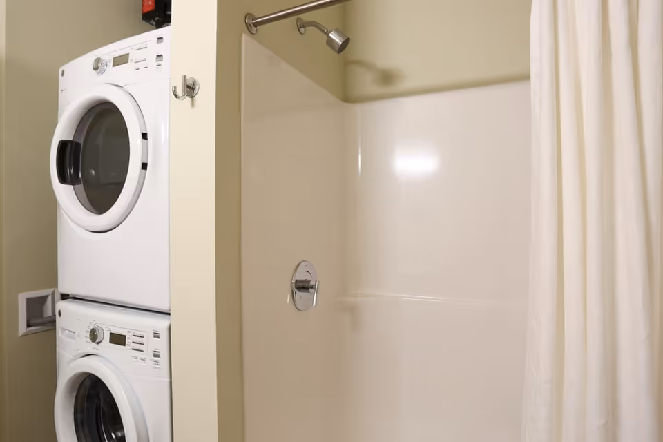 A small laundry area with a stacked white washer and dryer next to a shower with a white curtain and a silver showerhead.